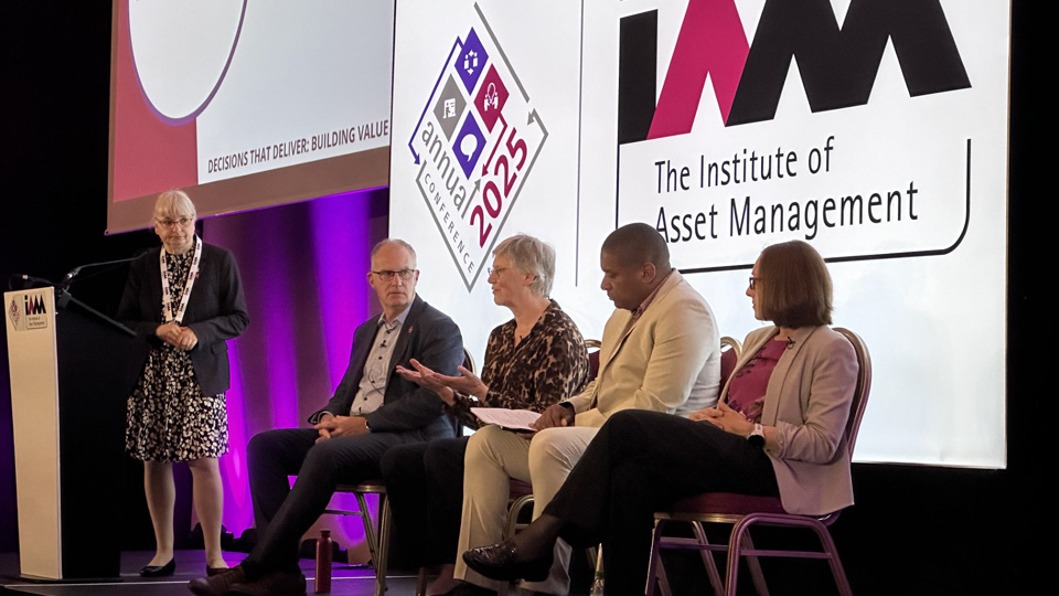 Five people on stage at an IAM event; one stands at a podium with the IAM logo, and a screen behind displays 